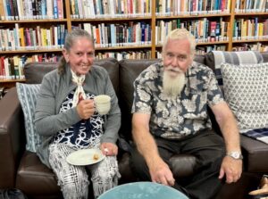 Two members, man and woman, of the Laryngectomee Association of NSW, seated on a sofa, smiling and facing the camera. The lady is holding a cup of tea.