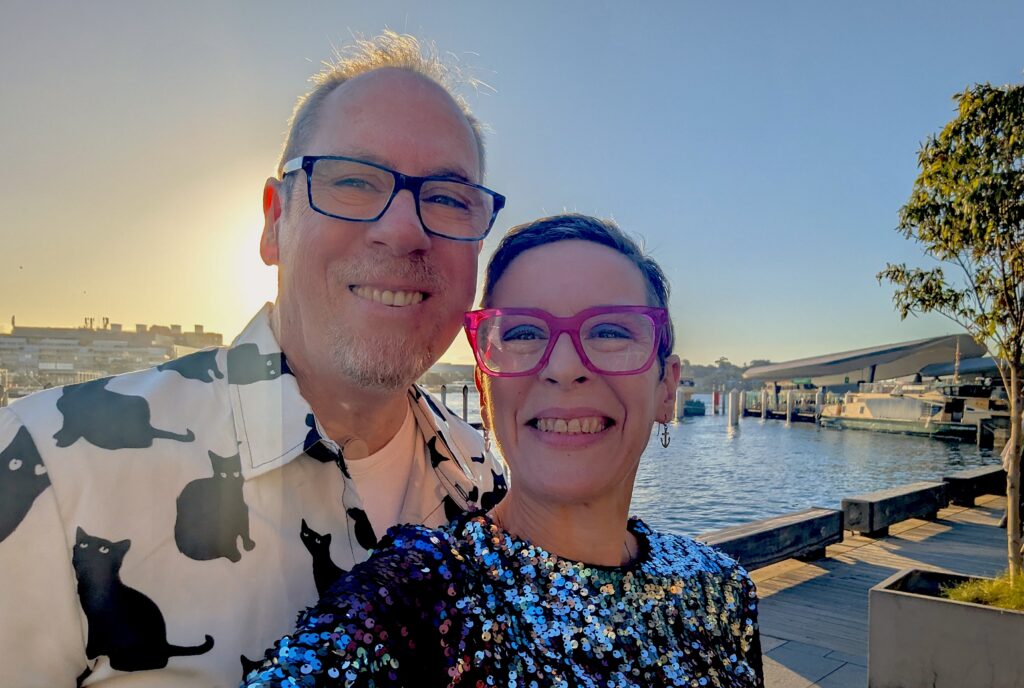 had and shoulder photo of male and female, both wearing glasses, standing in front of waterfront with boats on water