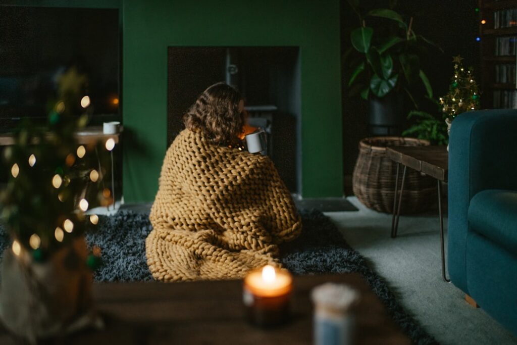 image of female wrapped in a beige blanket sipping a beverage from a mug, in front of a fire stove.