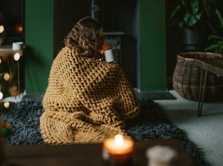 image of female wrapped in a beige blanket sipping a beverage from a mug, in front of a fire stove.